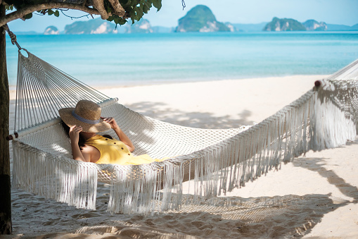 Happy Woman Traveler is relaxing in a hammock on the paradise beach. Female tourist in yellow dress rest near tropical sea. vacation, travel, summer, Wanderlust and holiday concept