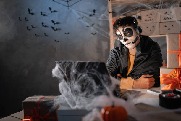 el dueño de un negocio hombre árabe usa calavera de arcilla blanca, maquillaje profesional en la oficina en casa con auriculares. sentado aburrido. oficina decorada festivamente. concepto de venta de halloween. estandarte. - freight transportation audio fotografías e imágenes de stock