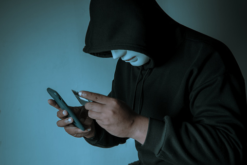 An anonymous masked hacker is using a smartphone to penetrate credit card financial information. Hacking and malware concept.