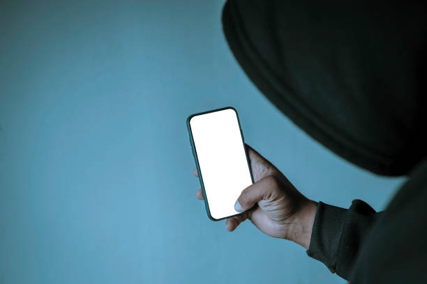un hacker anonimo che indossa una maschera e una felpa nera con cappuccio mostra uno smartphone con uno schermo bianco vuoto. concetti di hacking e malware. - business stealing top secret confidential foto e immagini stock