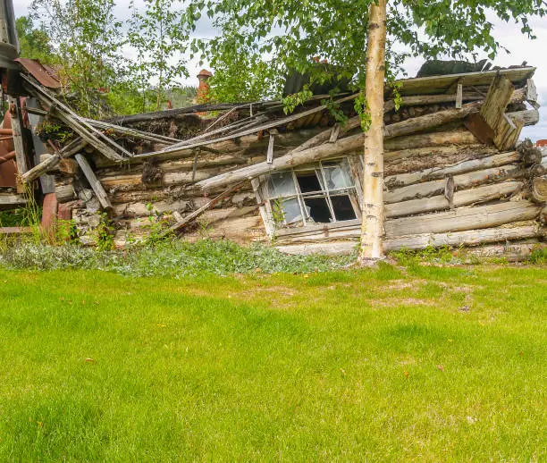 Remains of old log cabin with broken window falling down in green field.