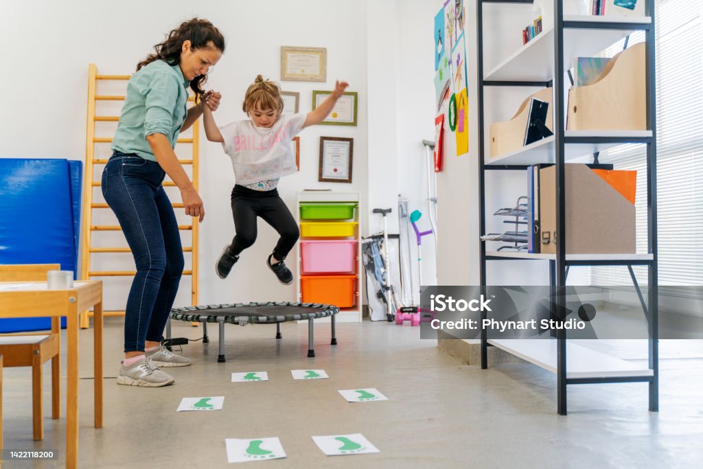 Down Syndrome: Occupational Therapy girl with down syndrome jumps on trampoline with the help of trainer Child Stock Photo