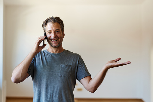 Happy man talking on mobile phone while holding his palm open and looking at camera in his new apartment. Copy space.