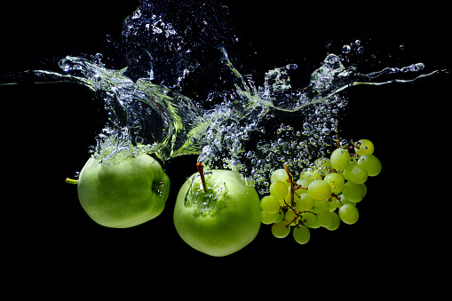 Green apples and white grapes dropped in water with splashes and bubbles isolated on black background.