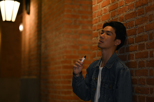 Man smoking with electronic cigarette standing in the night city with blurred night street lights background.