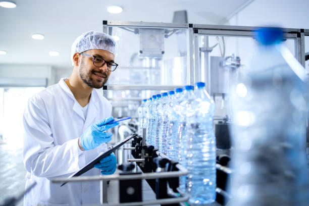 istock Technologist working in bottling factory doing quality control of drinking water before distribution. In background factory interior and industrial conveyor machine transporting PET bottles with water. 1422138228