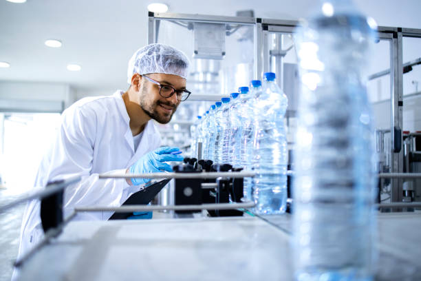 istock Technologist working in bottling factory doing quality control of drinking water before distribution. In background factory interior and industrial conveyor machine transporting PET bottles with water. 1422139432