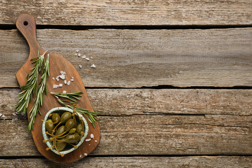 Delicious pickled capers and rosemary twigs on wooden table, top view. Space for text