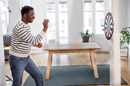 A handsome African-American man is throwing darts at the dartboard at home.