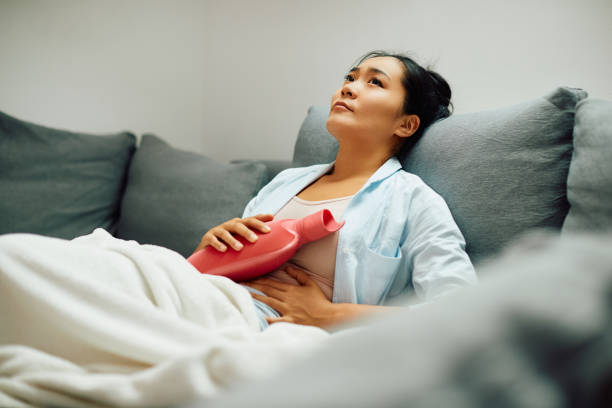 Young Asian woman holding a hot water bottle on her stomach at home. Young Asian woman relieves stomach pain with hot water bottle while relaxing on sofa at home. endometriosis stock pictures, royalty-free photos & images