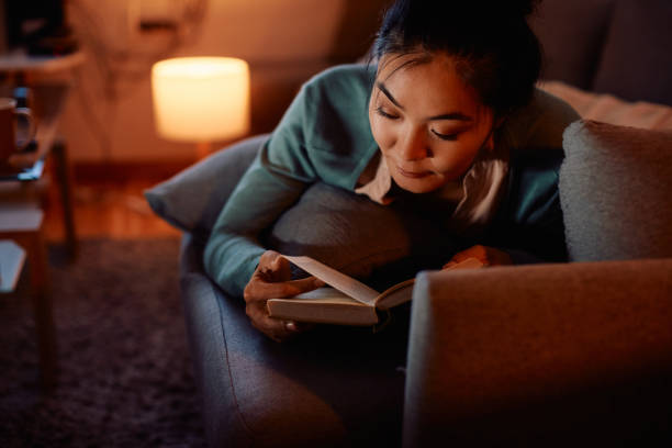 istock Young Asian woman reading book while relaxing on the sofa in the evening. 1422145008