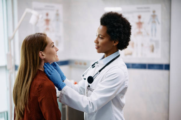 Smiling woman having a thyroid exam by black female endocrinologist at medical clinic. African American female doctor touching woman's neck during thyroid exam at medical clinic. visit stock pictures, royalty-free photos & images