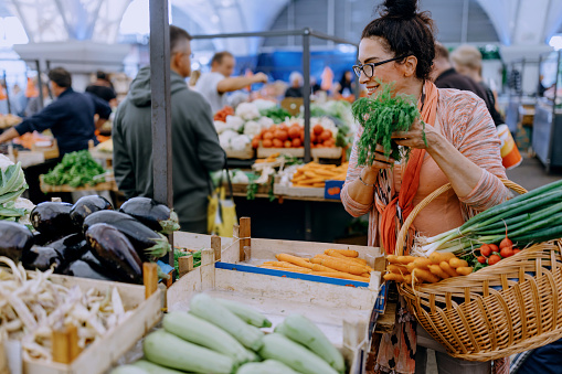 A mature Caucasian woman is spending time shopping at the farmer's market, holding a basket and carrots with a smile on her face.