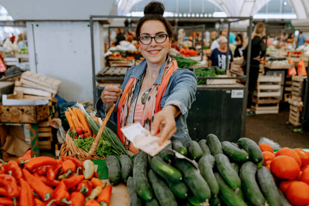 A mature Caucasian woman is looking at the camera with a smile on her face, while paying for her bill at the farmer's market.