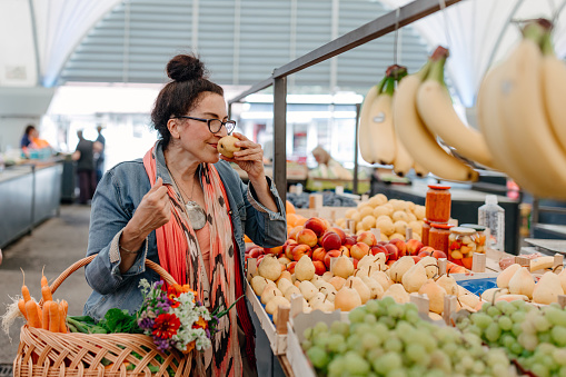 A mature Caucasian woman is shopping for fresh fruit at the farmer's market, stopping to smell a pear with her eyes closed.