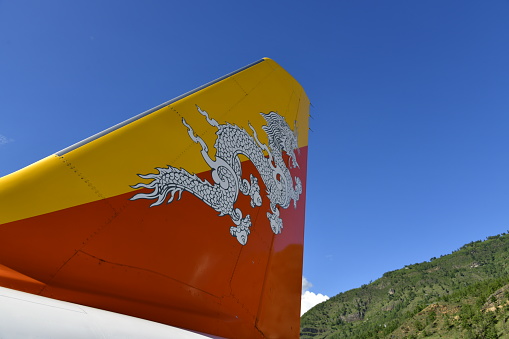 Paro, Bhutan: flag of Bhutan on an aircraft tail - The two colors of the flag, divided diagonally, represent spiritual and temporal power within Bhutan. The orange part of the flag represents the Drukpas monasteries and Buddhist religious practice, while the saffron yellow field denotes the secular authority of the dynasty. The dragon ('Druk'), is the Tibetan name for the kingdom of Bhutan. The jewels clamped in the dragon's claws symbolize wealth. The snarling mouth represents the strength of the male and female deities protecting the country.