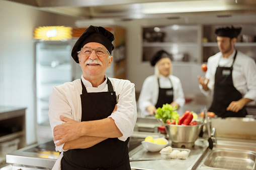 Portrait of senior chef standing in commercial kitchen at restaurant with his colleagues in the background