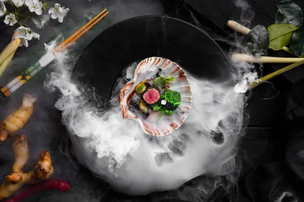 Roll with tuna, nori, gold sprinkle, chuka, tobiko caviar, sesame seeds, parsley and hazelnuts in a shell in a plate of smoke on a black table with a cloth, ginger, chopsticks, a cherry sprig and rose leaves