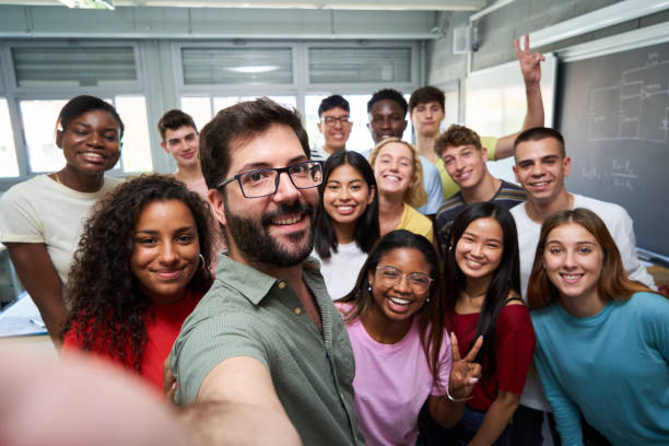 Happy selfie of young group of Erasmus students taking a photo with their teacher in the classroom. Happy selfie of young group of students taking a photo with their male teacher in the classroom, celebrating the end of course. Classmates from different countries, looking at camera with big smiles. education student mobile phone university stock pictures, royalty-free photos & images