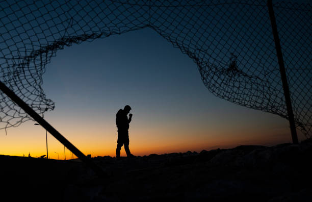 Refugee man standing behind the fence at sunrise stock photo