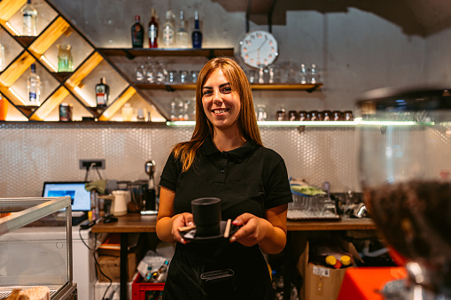 Beautiful young smiling female barista behind the bar counter, holding a cup of coffee.