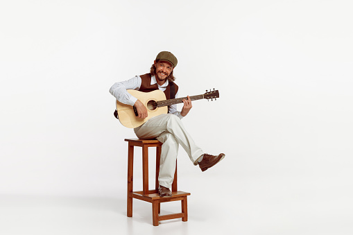 Portrait of stylish man playing guitar, performing isolated over white studio background. Smiling. Cheerful song. Concept of live music, performance, retro style, creativity, artistic lifestyle