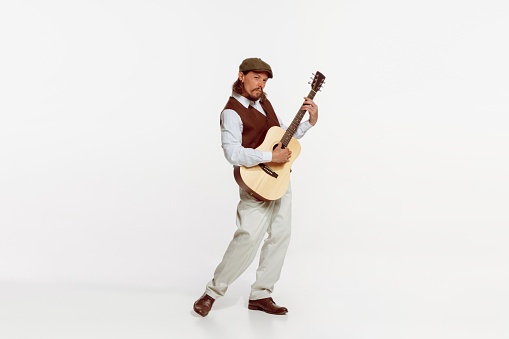 Portrait of stylish man playing guitar, performing isolated over white studio background. Making lovely sounds. Concept of live music, performance, retro style, creativity, artistic lifestyle