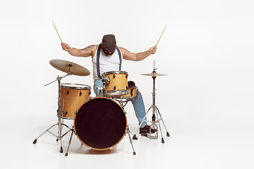Portrait of emotive, expressive man in sunglasses playing drums, performing isolated on white background. Rock concert. Concept of live music, performance, retro style, creativity, artistic lifestyle