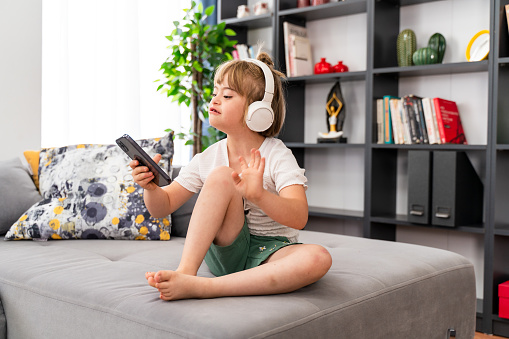 Portrait of girl with Down syndrome using smartphone. Diversity kid listening to music in headphones sitting on sofa at home. Audio development for children with disabilities, inclusive education