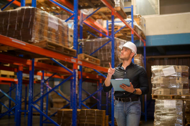 istock Young worker wearing helmet checking inventory and counting product on shelf in modern warehouse. 1422203599