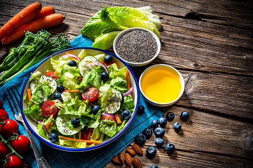 High angle view of a healthy green vegetables salad shot on rustic wooden table background. Bowls with chia seeds, olive oil, almonds and cherry tomatoes complete the composition. High resolution 42Mp studio digital capture taken with SONY A7rII and Zeiss Batis 40mm F2.0 CF lens