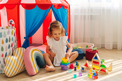 A little girl sitting on floor plays with colorful toys in playroom. Educational game for baby and toddler in modern nursery.