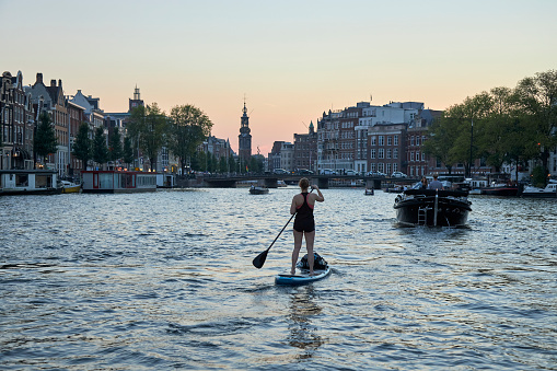 Woman paddleboarding with The Mint Tower in the background at twilight, The Amstel River, Amsterdam, The Netherlands.