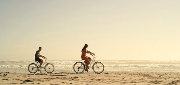Beach, romance and a couple cycling by the sea, romantic ride during sunset or sunrise. Love, travel and relax in nature, calm bicycle cruise for man and woman. Waves, sand and the sky in background.