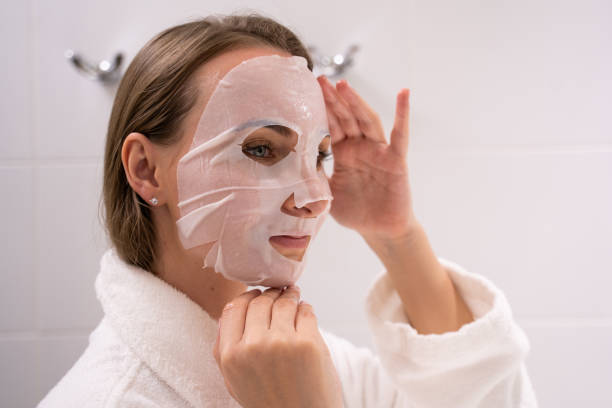 istock Woman in robe puts sheet mask on face adjusting with fingers 1422223081