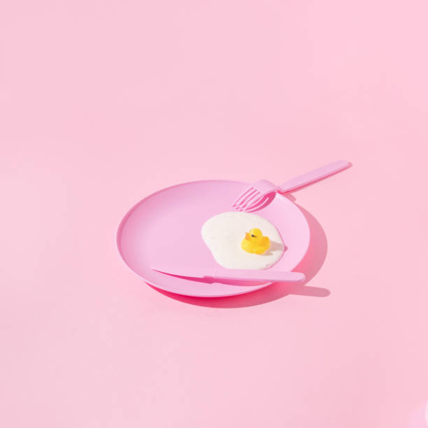 istock Creative layout with pastel pink plate, fork, knife, yellow rubber duck and white paint on pastel pink background. Aesthetic fashion food restaurant concept. Minimal surreal breakfast  idea. 1422225597