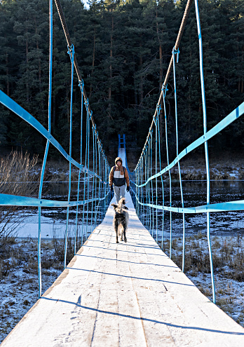 Young man with mixed breed fluffy dog playing and walking on wooden bridge over river covered with first snow and ice in autumn or early winter Hiking and traveling with pets Active lifestyle dog adoption