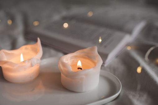 Burning scented candles on white ceramic tray with open paper book in bed close up. Cozy hygge home atmosphere. Winter war season.