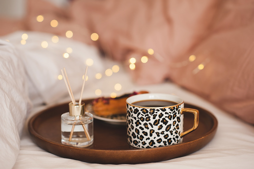 Cup of coffee in tray with home fragrance in glass bottle with sticks and tasty cake in bed close up. Good morning. Breakfast time. Aromatherapy. Home cozy atmosphere.