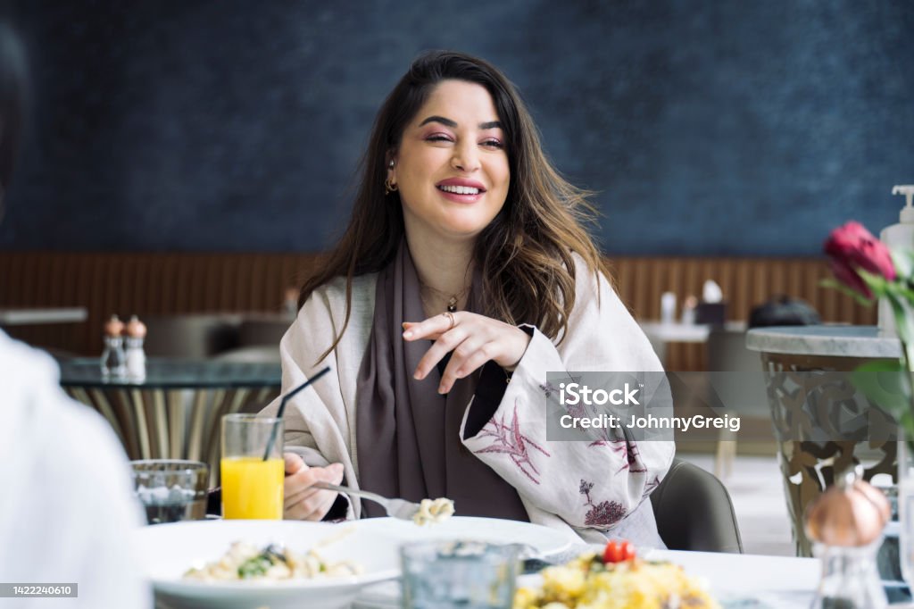 Cheerful Saudi woman dining at restaurant with friends Candid portrait of longhaired Middle Eastern woman in late 20s wearing abaya and laughing while enjoying light lunch and conversation with friends. Restaurant Stock Photo