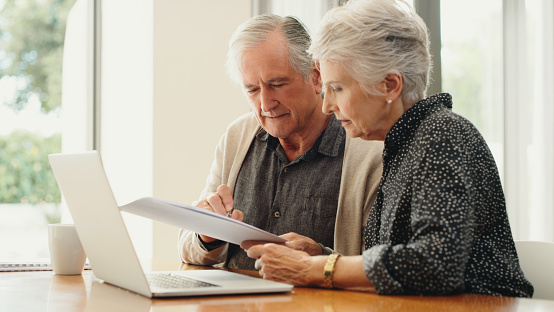 Senior couple with laptop and budget document, paperwork or finances for retirement plan or pension expenses or mortgage home loan. Married man and woman managing utility bills or debt online
