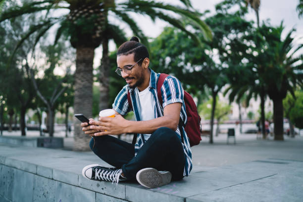 istock Happy young black hipster male with backpack using smartphone while sitting on step and enjoying takeaway coffee 1422242705