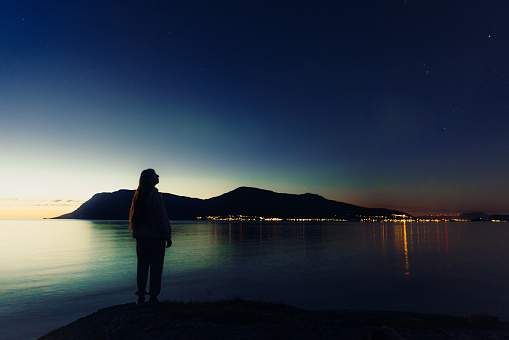 Silhouette of female admiring Aurora Borealis in the background with the mountains and the city in lights, More og Romsdal county, Norway