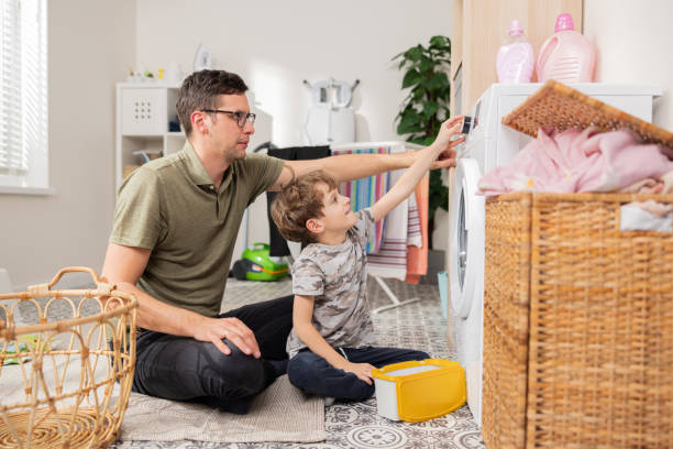 vater verbringt zeit mit kleinen kindern in der waschküche, im badezimmer, erledigt während des mutterschaftsurlaubs haushaltsaufgaben, kümmert sich um den sohn, zeigt dem jungen, wie man eine waschmaschine bedient - laundry husband housework men stock-fotos und bilder