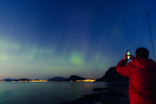 One male in red jacket taking photo of of the strong Aurora Borealis with the mountains and the city in light, More og Romsdal county, Norway
