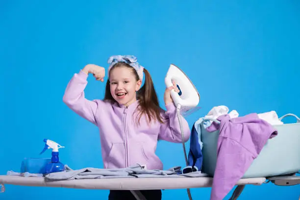 Little smiling sweet girl irons clothes with iron, strongwoman raises hand to show biceps, child defeated a mountain of laundry items.