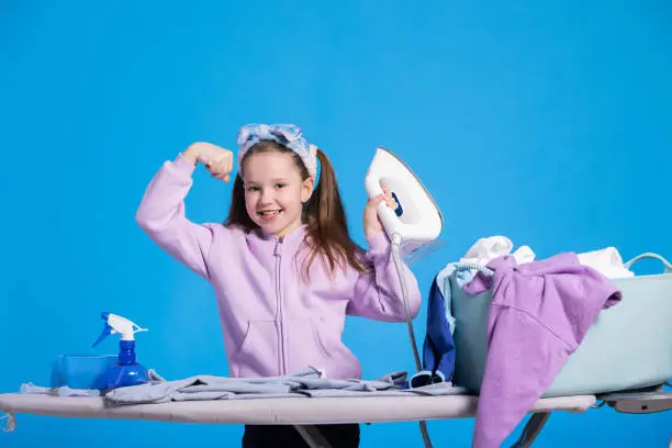 Little smiling sweet girl irons clothes with iron, strongwoman raises hand to show biceps, child defeated a mountain of laundry items.