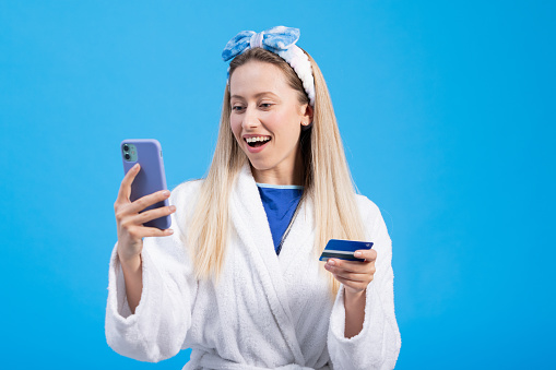 A young woman in a bathrobe poses on a blue studio background holding a credit card and a phone in hand, places an order in an online store for cosmetics.