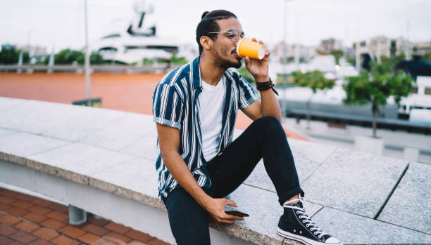 istock Positive dark skinned male traveler resting on urban setting drinking beverage from disposable cup, 20s handsome teen african american hipster guy recreating outdoors in city during weekends 1422253083