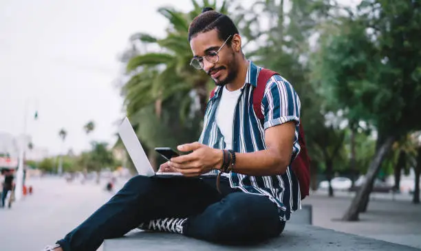 Young millennial dark skinned guy holding mobile phone reading information about online booking, hipster guys spending time time outdoors holding mobile phone working via laptop on freelance